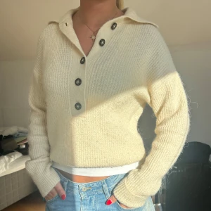 Beige stickad tröja med knappar - Supermysig beige stickad tröja med lång ärm och snygg krage. Tröjan har en rak passform och coola svarta knappar framtill som detalj. Perfekt att styla med jeans för en chill look.