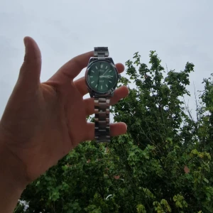 Grön klocka med metallarmband - Stilren klocka från Olevs med grön urtavla och datumvisning. Klockan har ett klassiskt runt boett i silverfärgad metall och matchande länkarmband. Perfekt accessoar för dig som vill sticka ut med en cool färg på handleden.