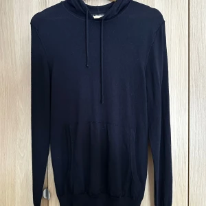 Marinblå hoodie från J.Lindeberg - Marinblå hoodie från J.Lindeberg i storlek L men lite krympt i tvätten sä skulle säga storlek M. Tröjan har en klassisk huva med snörning, långärmade ärmar och en stor magficka framtill. Tillverkad i mjukt stickat material som känns skönt mot huden. Perfekt för en stilren och avslappnad look. 