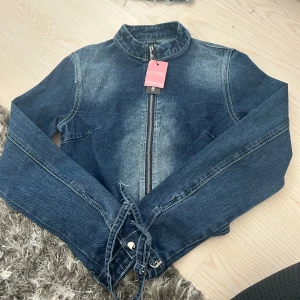 Jeansjacka från PrettyLittleThing med spännen och matchande kjol  - Snygg blå jeansjacka med dragkedja framtill och coola snörningsdetaljer vid ärmsluten. Jackan har en rak passform och klassisk krage. Perfekt för dig som vill ha en trendig denimlook med extra edge.