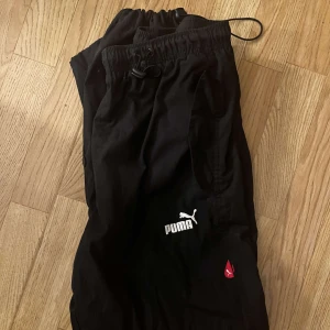 Svarta vindbyxor från Puma - Svarta vindbyxor från Puma med elastisk midja och snörning. Byxorna har två sidofickor och en vit Puma-logga på benet samt en liten röd detalj.🩷