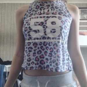 Leopardmönstrad croppad topp H&M - Säljer en croppad ärmlös topp från H&M med leopardmönster i blått, rosa och svart. Framsidan har trycket 'HAWAII 58' och baksidan är öppen med twistad omlott-design. Perfekt för dig som vill sticka ut med sportig vibe och coolt mönster.