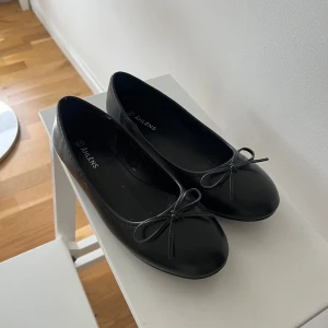 Svarta ballerinaskor från Åhléns - Klassiska svarta ballerinaskor från Åhléns med rund tå och en söt rosettdetalj framtill. Skorna har platt sula och är tillverkade i syntetmaterial, vilket gör dem lätta och smidiga att bära. Perfekta för en stilren och enkel look.
