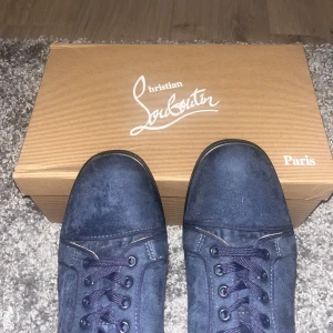 Mörkblå mocka sneakers från Louboutin - Snygga mörkblå sneakers från Christian Louboutin i mjuk mocka med klassisk röd sula och svarta detaljer. Skorna har rund tå, snörning och diskret logga på sidan. Insidan är ljusbeige och sulan har den ikoniska Louboutin-signaturen.