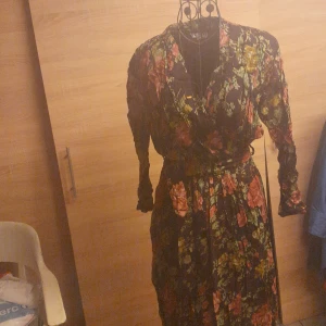 Blommig långklänning från Together - Långklänning från Together i storlek 36 med svart bas och färgglatt blommönster i grönt, rött och gult. Klänningen har långa ärmar, omlottliv och knytband i midjan för en snygg siluett. Perfekt för dig som gillar romantiska vibes och boho-stil.