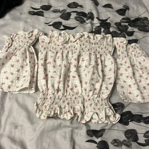 Vit blommig offshoulder blus - Supersöt vit blus med små röda blommor och gröna blad, offshoulder och långa puffiga ärmar. Smockad överdel och volangkant nertill ger en romantisk vibe. Perfekt för vår och sommar när du vill känna dig extra gullig.