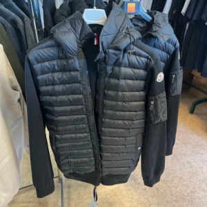 Svart dunväst med huva från Moncler - Snygg svart dunväst från Moncler med quiltad framsida och huva. Västen har dragkedja, fickor med dragkedja på ärmen och klassisk Moncler-logga. Materialmix med vadderad front och ärmar i ull för en modern look.