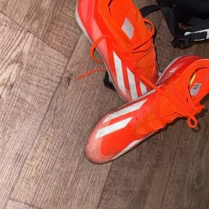 Röda Adidas fotbollsskor med snörning - Snygga röda fotbollsskor från Adidas med vita detaljer och klassiska ränder på sidorna. Skorna har snörning och en smidig, sportig design som passar perfekt för planen. Tillverkade i syntetmaterial för lätt känsla och bra grepp. Jag kan tvätta dom innan de skickas så de blir som nya.använd 2 gånger. Storlek 43