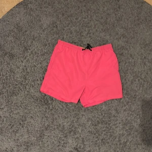 Rosa badshorts med snörning - Snygga rosa badshorts med elastisk midja och svart snörning framtill. Shortsen har en bakficka med liten ögla och är tillverkade i ett lätt syntetmaterial som torkar snabbt. Perfekta för stranden eller poolen i sommar.