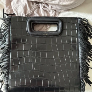 Svart handväska med fransar från Maje - Säljer en svart handväska från Maje i croco-präglat skinn med rektangulära handtag och coola fransar längs sidorna. Långt band och dustbag medföljer!