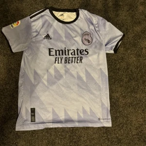 Real Madrid Benzema Adidas fotbollströja - Snygg Real Madrid fotbollströja från Adidas med Benzema och nummer 9 på ryggen. Tröjan är ljuslila med geometriskt mönster, svarta detaljer och klubbmärke på bröstet. Korta ärmar och tillverkad i lätt, ventilerande material. Perfekt för alla Madridistas!