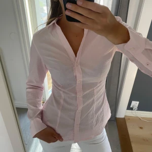 Rosa- och vitrandig skjorta från H&M - Säljer en figursydd skjorta från H&M i storlek 36. Skjortan är ljusrosa med tunna vita ränder, har lång ärm och klassisk krage. Knappar framtill och tillverkad i bomull. Perfekt för en clean och fräsch look.