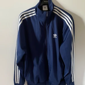 Blå Adidas track jacket med vita ränder - Klassisk marinblå Adidas track jacket med vita ränder längs ärmarna och broderad logga på bröstet. Jackan har dragkedja framtill, hög krage och ribbade muddar. Perfekt för en sportig och avslappnad stil. Pris kan diskuteras.