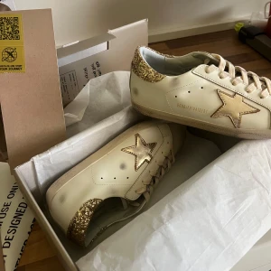 Golden Goose Superstar sneakers guldstjärna - Snygga Golden Goose Superstar sneakers i beige skinn med guldglittrig häl och metallic guldstjärna på sidan. Klassisk rund tå, platt sula och beige snörning. Perfekta för dig som gillar unika detaljer och lyxig streetstyle. Storlek 40, använt 1-2 gånger. Kvitto finns. Skriv för mer bilder eller vid frågor!🌸