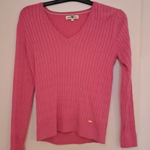 Rosa stickad tröja Hampton Republic - Supersnygg rosa stickad tröja från Hampton Republic med klassiskt kabelstickat mönster och v-ringning. Långärmad modell med ribbade muddar och liten guldfärgad logodetalj nertill. Perfekt för dig som gillar färg och stilren look.