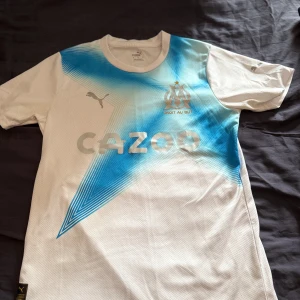 Marseille matchtröja Puma M - Säljer en officiell Olympique de Marseille matchtröja från Puma i storlek M. Tröjan är vit med blått grafiskt mönster över bröstet, klubbmärke och Puma-logga i silver samt sponsortryck. Tillverkad i lätt och ventilerande polyester, perfekt för fotboll eller träning.