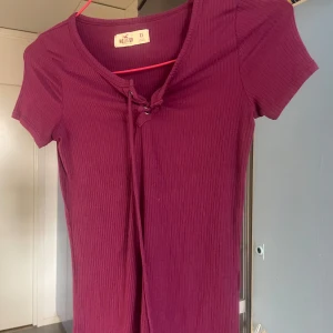 Ribbad vinröd top Holister  XS - Supersnygg vinröd ribbad top från Hollister med kort ärm och snörning vid halsen. Figurnära passform och mjukt material som känns skönt mot huden. Perfekt för dig som gillar en enkel men trendig stil.