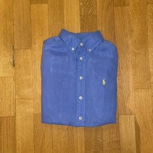 Blå linneskjorta från Polo Ralph Lauren - Snygg blå skjorta från Polo Ralph Lauren i linne med klassisk krage och knäppning framtill. Skjortan har långa ärmar och den ikoniska gula broderade loggan på bröstet. Perfekt för en chill och stilren look.
