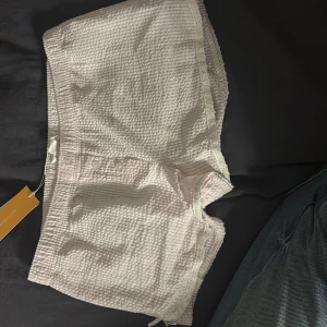 Ljusrosa shorts med spets och rosett - Supergulliga ljusrosa shorts i bomull med våfflad struktur, spetskant och liten rosett på benet. De har elastisk midja, knappar framtill och en söt, feminin vibe. Perfekta för varma dagar eller som mysplagg hemma.