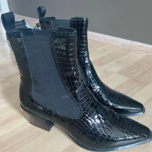 Svarta spetsiga boots - Snygga svarta chelsea boots med glansig croco-präglad yta och spetsig tå. Skorna har bred resår på sidorna och en stadig klack som ger extra attityd.