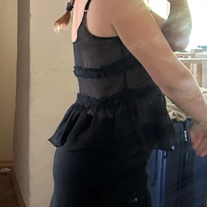 Svart knytt med volang från ONLY - Supersöt svart omlottopp från ONLY i transparent, lätt material. Toppen har smala axelband, volangdetaljer och knyt framtill. Den är figurnära upptill och har en lösare, volangig nederdel som ger en romantisk vibe.