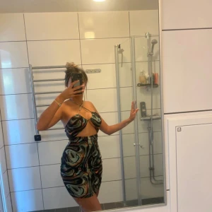 Mönstrad kort klänning med cut-out - Säljer en supercool kort klänning med swirl-mönster i svart, beige, grön och brun. Klänningen är axelbandslös med cut-out framtill och har en tight passform som framhäver figuren. Perfekt för dig som vill sticka ut!