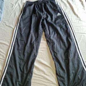 Svarta Adidas träningsbyxor med ränder - Svarta träningsbyxor från Adidas med klassiska vita ränder längs sidorna och Adidas-logga på låret. Passar för både tjejer och killar.