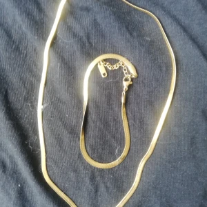 Guldigt halsband och armband set - Snyggt set med ett guldigt halsband och matchande armband i platt, ormliknande design. Båda har en stilren och minimalistisk look som passar perfekt till din outfit. Lätt att justera med smidig kedja och lås. Ej äkta guld 