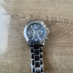 Silverfärgad klocka från Boss - Stilren klocka från Boss med silverfärgat armband i metall och blå urtavla. Klockan har tre mindre urtavlor, markerade siffror på ringen och robust länkarmband. Perfekt accessoar för dig som gillar klassisk design med sportig känsla.