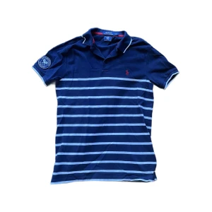 Ralph Lauren wimbeldon piké  - Snygg blå pikétröja från Polo Ralph Lauren med vita horisontella ränder och röd broderad logga på bröstet. Klassisk krage med knappslå och vit piping. Tillverkad i bomull, perfekt för en stilren och sportig look. Jag skickar ej bild när jag bär plaggen och jag vet ej vilken längd och vikt de passar då jag inte prövar kläderna jag säljer 😃