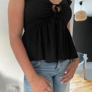 Svart blus med knyt från 157 - Supersöt svart blus från 157 med knyt framtill och liten cutout vid bysten. Modellen är luftig med volangliknande nederdel och ärmlös design. Perfekt till jeans eller kjol för en chill och trendig look.