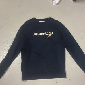 mörkblå sweatshirt från Axel Arigato - Svart sweatshirt från Axel Arigato med trycket 'ARIGATO ©2014' och en liten fågel på bröstet. Klassisk rund halsringning och långa ärmar. Mjuk insida och ribbade muddar vid ärmslut och nederkant. Perfekt för en clean och stilren look.