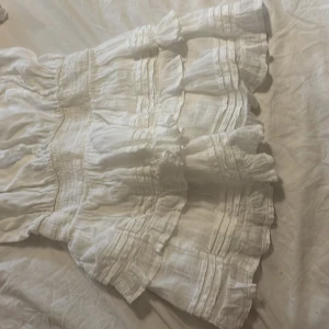 Vit volangklänning från Gina Tricot - Superfin vit kortklänning från Gina Tricot med volanger och smock i midjan. Klänningen har korta ärmar, v-ringning i ryggen och är gjord i ett lätt bomullstyg. Perfekt för sommarens alla tillfällen och ger en romantisk vibe.