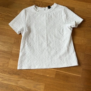 Vit strukturerad blus från Geena - Säljer en vit blus från Geena med korta ärmar och rund hals. Tyget har ett snyggt strukturerat mönster som ger en cool 3D-effekt. Blusen har dragkedja i ryggen för enkel påklädning. Perfekt för dig som gillar stilrena och unika plagg.