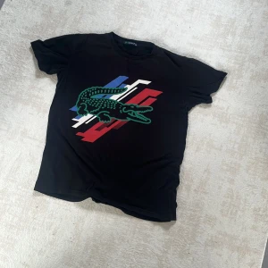 Svart Lacoste t-shirt med tryck - Svart t-shirt från Lacoste med stort grönt krokodiltryck och grafiska detaljer i blått, vitt och rött på bröstet. Klassisk rund hals och korta ärmar. 