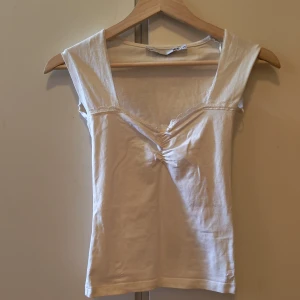 Vit linnetopp med spets från 157 - Supersöt vit linnetopp från 157 i storlek XS. Toppen har v-ringning med spetskant och en liten rosett framtill. Den är ärmlös och har en lätt rynkning vid bysten som ger en snygg form. Perfekt för en fräsch och enkel look.