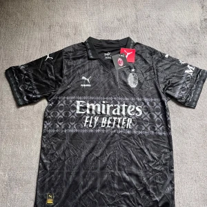 Svart AC Milan x Puma fotbollströja - AC Milan x Puma fotbollströja i svart med unikt mönster och silverfärgade loggor. Tröjan har korta ärmar, krage och tryck från Emirates och wefox. Materialet är lätt och andas, perfekt för match eller träning. Snygg detaljrik design med klubbmärke på bröstet.