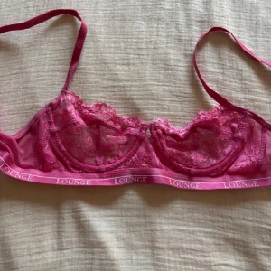 Rosa spetsig bralette från Lounge - Säljer en snygg rosa bralette från Lounge med spetsdetaljer och tunna axelband. Underkanten har en elastisk resår med Lounge-loggan tryckt runtom. Perfekt för dig som gillar en feminin och bekväm stil. Liten u storleken så sitter mer som 70c typ💕