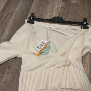 Krämvit omlottkofta från Gina Tricot - Superfin krämvit omlottkofta från Gina Tricot med bred ribbad kant nertill och knytband i sidan. Koftan har v-ringning och långa ärmar, perfekt för lager på lager. Mjuk och skön känsla, passar till allt och ger en clean look.