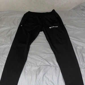 Svarta mjukisbyxor från Columbia M - Svarta mjukisbyxor från Columbia med elastisk midja och dragkedjeficka på sidan. Byxorna har en clean look med logga på låret och är tillverkade i mjukt syntetmaterial. Perfekta för chill eller träning.