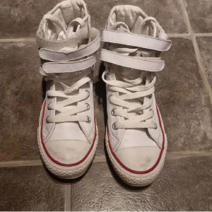Vita Converse All Star high-tops - Snygga vita Converse All Star high-tops i skinn med klassisk röd och blå rand runt sulan. Skorna har både snörning och två kardborreband uppe. Storlek 36.5 