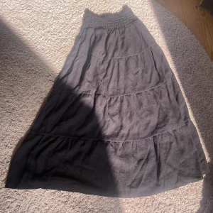 Svart långkjol från H&M - Svart långkjol från H&M med smockad midja och flera volanger. Kjolen är luftig och har en enkel, stilren look som passar perfekt till sneakers eller sandaler. Tillverkad i mjukt bomullstyg för en skön känsla hela dagen.