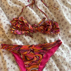 Färgglad bikini med volang och snörning - Snygg triangel bikini med färgstarkt  mönster i rosa, orange och gul. Överdelen har volangdetaljer och knyts i nacken. Bikinitrosan har matchande mönster och fodrad insida i rosa.  Knappt använt 