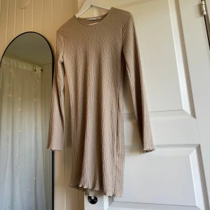Beige ribbad långärmad klänning - En enkel och stilren beige klänning. Modellen är kort, har lång ärm och är tillverkad i ett ribbat material som ger en snygg struktur. Perfekt för dig som gillar en clean och avslappnad look.
