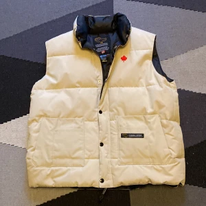 Beige dunväst från Canada Goose XL - Snygg beige dunväst från Canada Goose med svart insida och klassisk röd lönnlöv-detalj på bröstet. Västen har två stora fickor framtill, tryckknappar och hög krage. Yttermaterial i polyester och bomull, fodrad med ankdun för extra värme.
