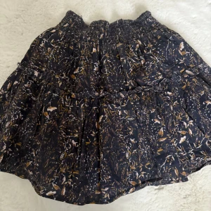 Svart blommig kortkjol med volang - Supersöt svart kortkjol med blommigt mönster i beige, vitt och grönt. Kjolen har bred resår i midjan och volangdetaljer som ger en flowy känsla. Perfekt för dig som gillar romantisk och bohemisk stil.