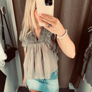 Neo noir blus - Supersöt grå blus med v-ringning och volangdetaljer längs axlarna. Blusen har en lös passform och är tillverkad i ett lätt, luftigt material som känns skönt mot huden. Perfekt att styla med jeanskjol eller shorts för en chill look.