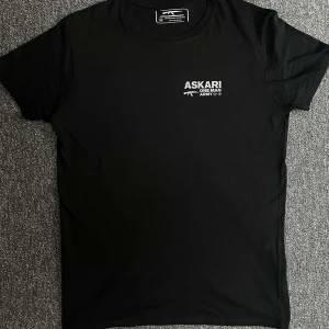 Svart ASKARI t-shirt med tryck - Svart t-shirt från Imaginations and Dreams med stort vitt ASKARI 'One Man Army'-tryck och gevärsgrafik på ryggen och mindre tryck på bröstet. Klassisk rund hals och korta ärmar. Skön passform och stilren look, perfekt för dig som gillar statement-plagg.