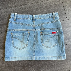 Ljusblå jeanskjol från Vero Moda - Snygg ljusblå jeanskjol från Vero Moda med klassisk femficksdesign, bälteshällor och knapp- samt dragkedjestängning. Kjolen har raka linjer och coola bakfickor med broderade detaljer och en liten röd logolapp. Perfekt för en avslappnad och trendig look.