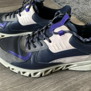 Ecco sneakers i blått och lila - Snygga sneakers från Ecco i en mix av mörkblått, grått och lila detaljer. Skorna har coola snörningar, robust sula med unik design och ovandel i skinn och syntet. Perfekta för dig som gillar sportig stil med edge. De är även vattentäta.